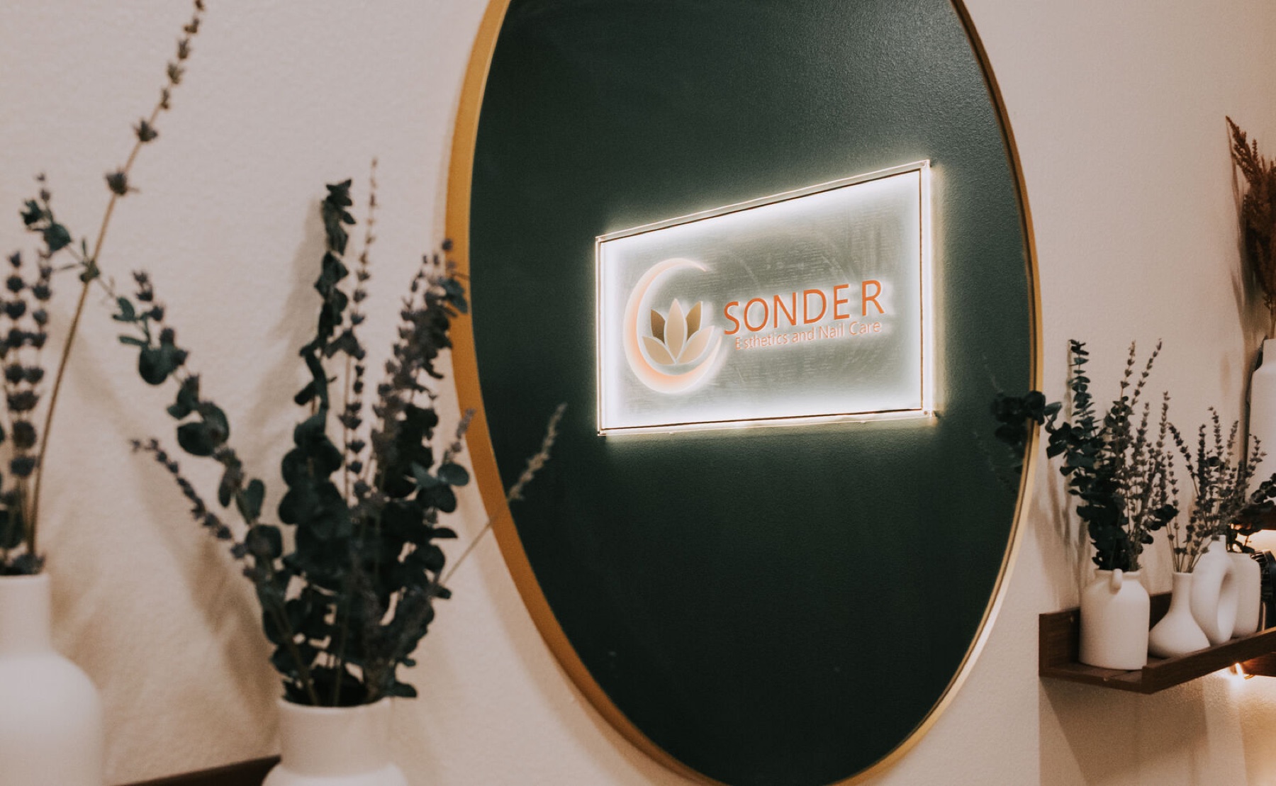 The Sonder Esthetics studio: the lit logo on a dark green wall, with eucalyptus